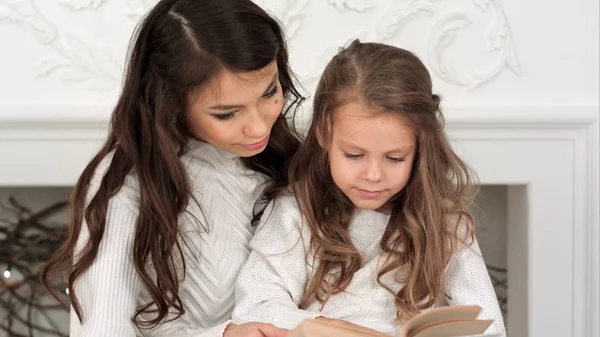 Happy mother and her beautiful daughter in white sweaters reading a book on Christmas eve — Stock Photo, Image