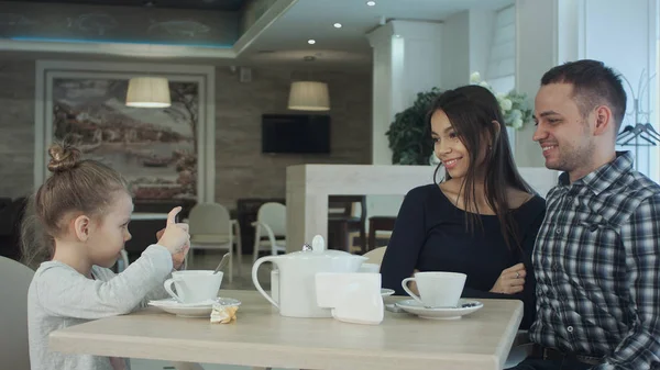 Mutlu anne, baba ve küçük kız cafede akşam yemeği. Çocuk alarak fotoğraf yanında smartphone kameraya poz veliler ise.