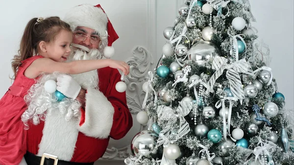 Fake Santa Claus decorating a Christmas tree holding a little girl in his arm