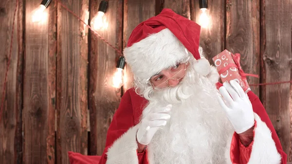 Santa Claus sacudiendo un pequeño regalo tratando de adivinar lo que hay dentro —  Fotos de Stock