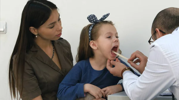 Pediatra masculino examinando la garganta de una niña sentada con su madre — Foto de Stock