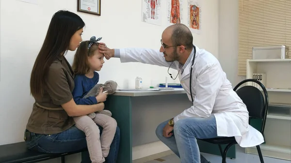 Pediatra masculino comprobando la temperatura de la cabeza de la niña con su mano y dándole termómetro — Foto de Stock