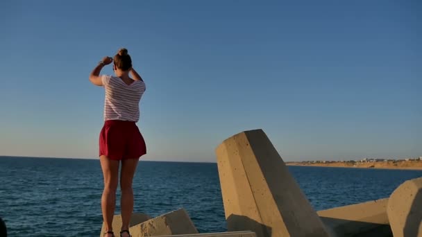 Красивая стройная девушка в шортах танцует на бетонных блоках у моря — стоковое видео