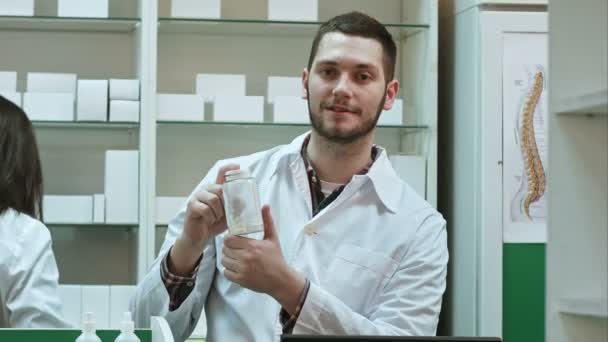 Joven malefarmacista sosteniendo un frasco blanco de pastillas, promoviendo la medicina, mientras su colega trabaja — Vídeo de stock