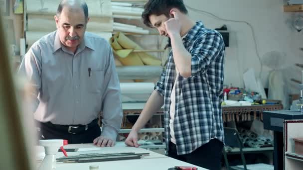 Старший работник-мужчина инструктирует молодого стажера, как построить каркас за столом в рамном цехе — стоковое видео