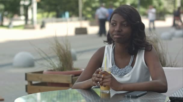 Retrato de una joven africana bebiendo jugo de naranja y soñando, mientras está sentada en la cafetería — Vídeo de stock