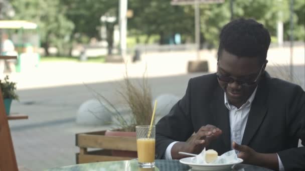Relajado despreocupado joven afroamericano hombre en gafas con estilo sentado solo en la mesa de la cafetería, el uso de teléfono inteligente, la lectura de mensajes de texto con sonrisa alegre durante el almuerzo — Vídeo de stock