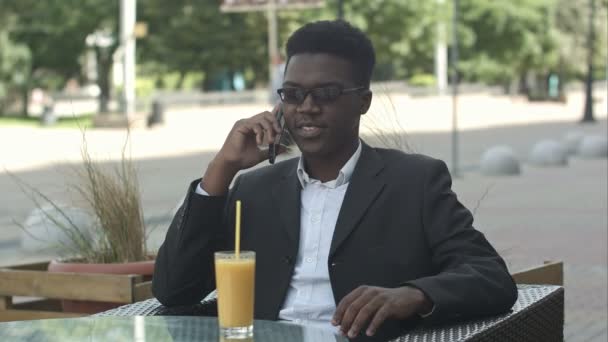 Experto en marketing afro-americano positivo en gafas y atuendo casual con llamada telefónica en cafetería — Vídeo de stock