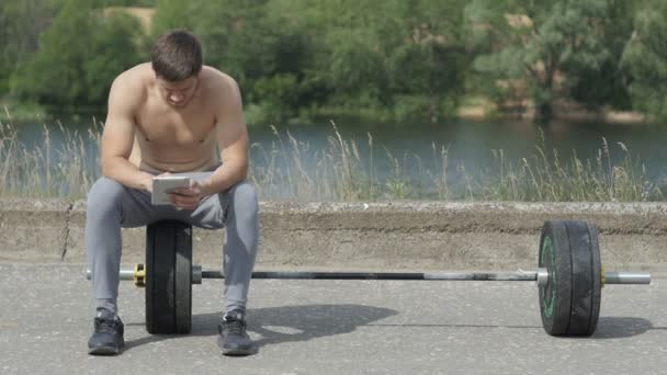 Молодий м'язистий чоловік сидить після фітнес-тренувань і використовує планшетний комп'ютер — стокове відео