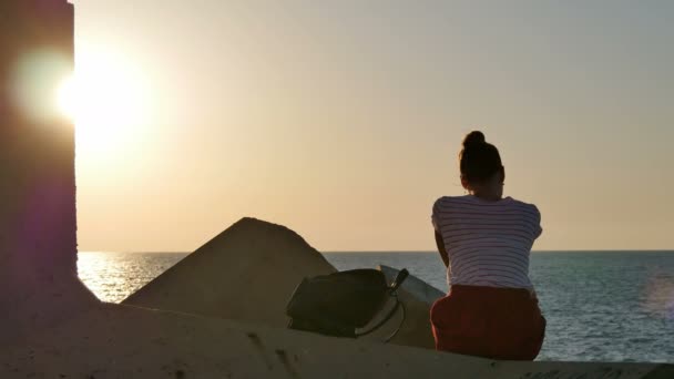 Мечтающая молодая женщина, сидящая на скалах у моря на закате — стоковое видео