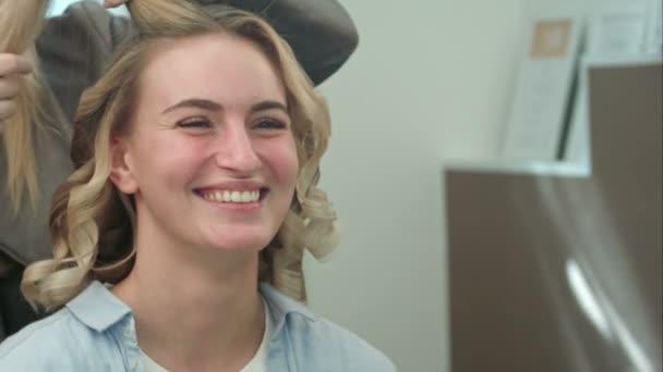Uma mulher em um salão de cabeleireiro esperando para ver os resultados de enrolar cachos, olhando em um espelho, sorrindo e conversando com o artista — Vídeo de Stock