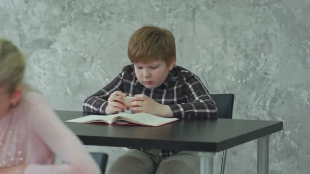 Молодая девушка в классе концентрируется на своем тесте, в то время как ее одноклассник играет в игры на смартфоне — стоковое видео