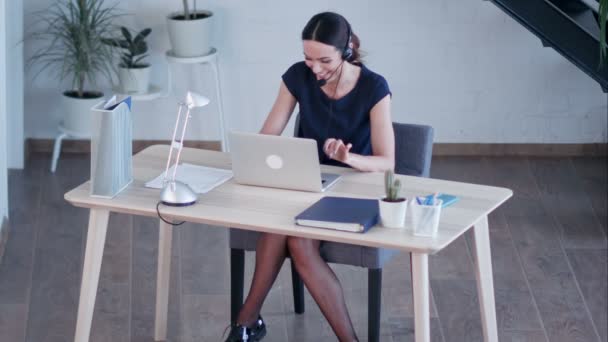 Gelukkig charmant jonge vrouw zitten en werken met laptop met behulp van headset in het kantoor — Stockvideo