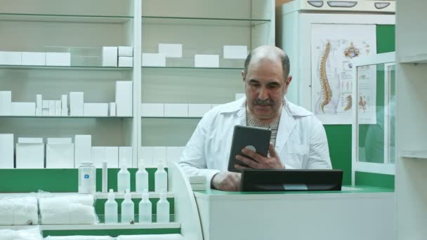 Старший фармацевт с усами работает на планшетном ПК, проверяя лекарства в аптеке — стоковое видео
