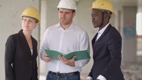 Groep van lachende bouwers in hardhats met Klembord en blauwdruk buitenshuis — Stockvideo