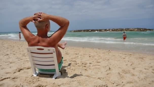 Zralý muž sedí na pláži židli a při pohledu na moře těší dovolená — Stock video