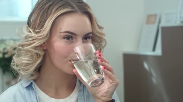 Прекрасная молодая женщина сохраняет здоровье, выпив стакан воды в помещении и улыбаясь — стоковое видео