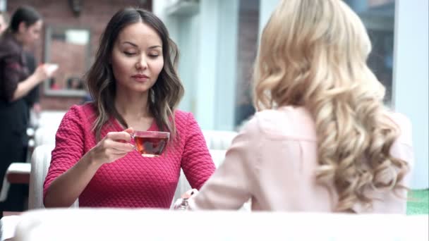Две молодые красивые девушки сидят в городском кафе с кофе и разговаривают — стоковое видео