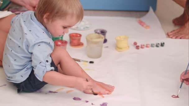 Lindo niño pequeño pintando sus pies en un gran papel blanco en blanco — Vídeos de Stock