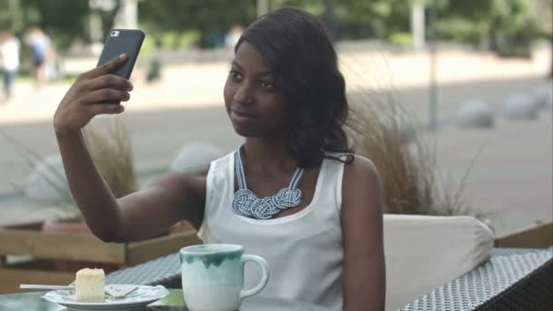 Atractiva joven africana sonriendo y tomando una selfie con su teléfono inteligente mientras está sentada sola en la cafetería al aire libre disfrutando de una comida — Vídeos de Stock