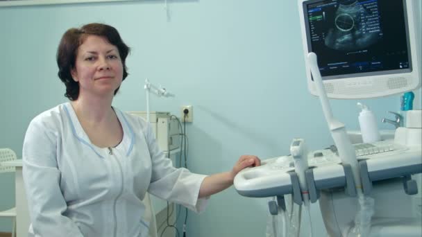 Усміхнена жінка-лікар сидить поруч з ультразвуковим діагностичним пристроєм — стокове відео