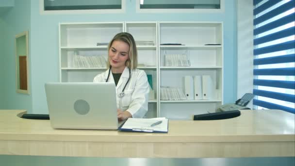 Улыбающаяся медсестра работает над ноутбуком и делает заметки на ресепшене — стоковое видео