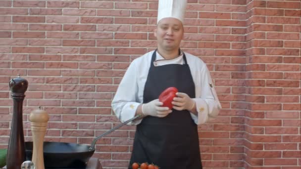 Middle-aged chef in white uniform standing near desk, talking to the camera with vegetable in his hands, ready to prepare a salad — Stock Video