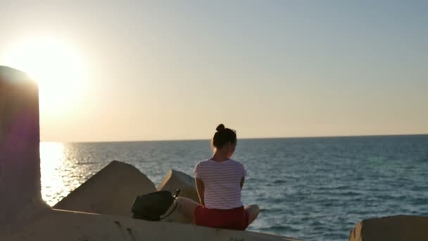 Drömmer unga kvinna som sitter på klipporna njuter av havet i solnedgången — Stockvideo