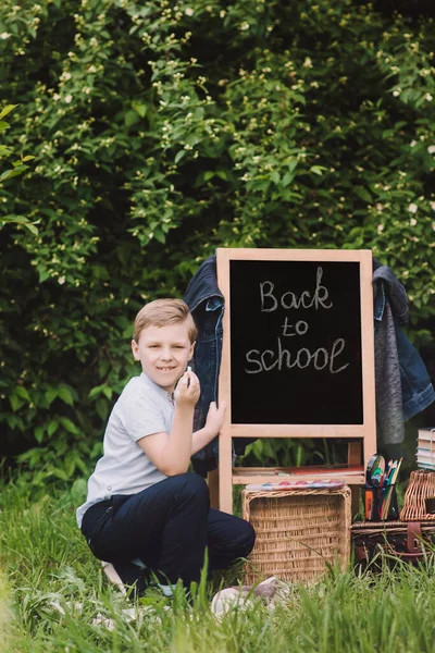 De vuelta a la escuela. Blonk feliz colegial escribe con tiza en pizarra. Lugar para el texto. Enfoque suave — Foto de Stock