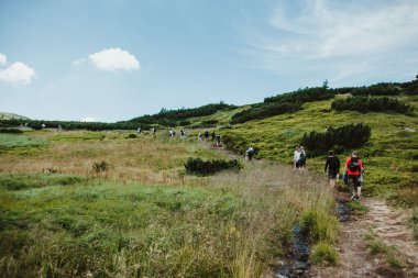 Dağ yolunda yürüyen sırt çantalı bir grup insanın siluetleri. Ufukta dağlar var. Gökyüzü mavi ve bulutlu.