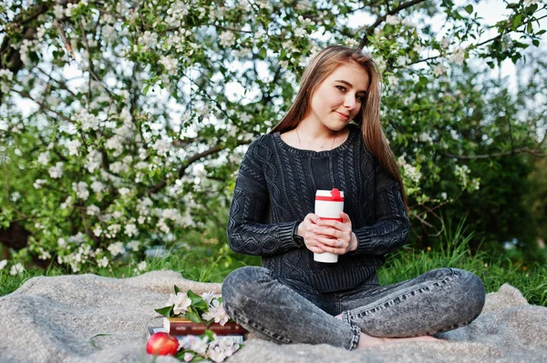 穿着牛仔裤的年轻黑发女孩坐在格子上对春天开花树和拿着茶热水瓶在手 — 图库照片