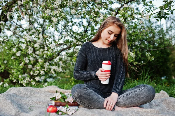 穿着牛仔裤的年轻黑发女孩坐在格子上对春天开花树和拿着茶热水瓶在手 — 图库照片