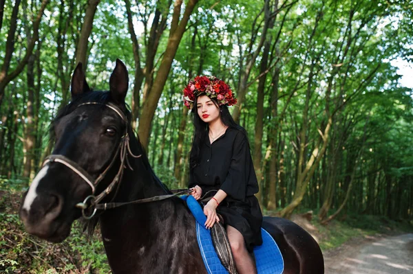 Mystical girl in wreath wear in black at horse in wood.