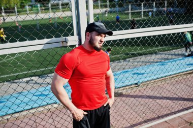 Genç acımasız sakallı kaslı adam giymek kırmızı gömlek, şort ve cap adlı Stadyumu.