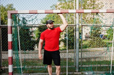 Genç acımasız sakallı kaslı adam giymek kırmızı gömlek, şort ve cap adlı Stadyumu.