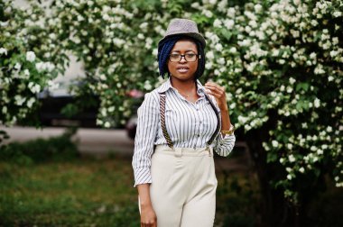 Şık Afro-Amerikan iş kadın pantolon askı ve bluz, şapka ve gözlük ile bloom çalılar karşı açık poz verdi.