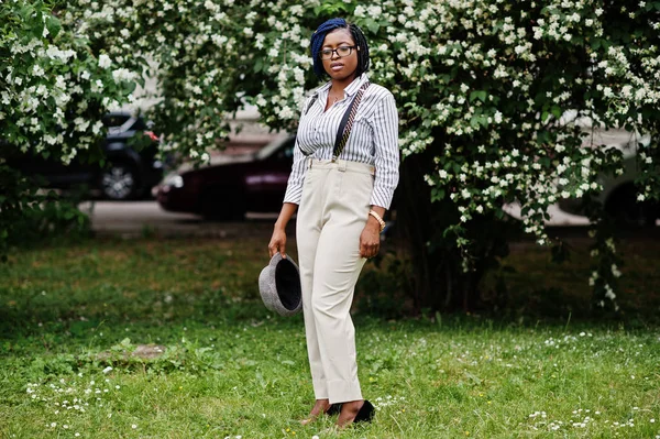 Şık Afro-Amerikan iş kadın pantolon askı ve bluz, şapka ve gözlük ile bloom çalılar karşı açık poz verdi.