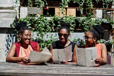 Güneşli yaz gün açık, elinde menüsüyle restoran masada oturan adlı üç şık Afrikalı-Amerikalı kadının poz.