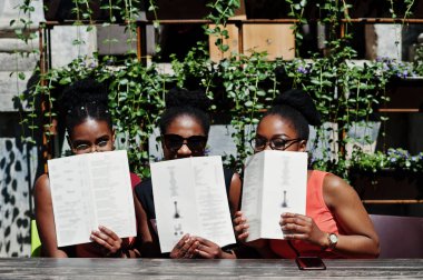 Güneşli yaz gün açık, elinde menüsüyle restoran masada oturan adlı üç şık Afrikalı-Amerikalı kadının poz.