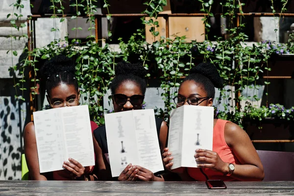 Güneşli yaz gün açık, elinde menüsüyle restoran masada oturan adlı üç şık Afrikalı-Amerikalı kadının poz.