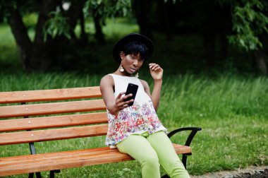 Yeşil pantolonlu, siyah şapkalı, parktaki bankta poz veren ve cep telefonuyla selfie çeken Afro-Amerikan manken kadın..