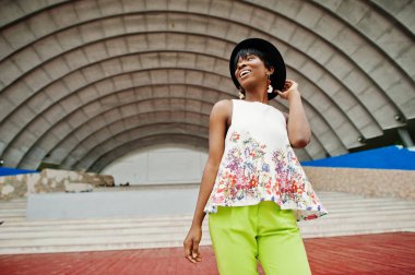 Afro-Amerikan model kadın yeşil pantolon ve siyah şapka şaşırtıcı arena hall karşı açık poz verdi.