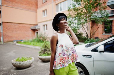 İnanılmaz bir Afro-Amerikan modeli kadın yeşil pantolon ve siyah şapka araba beyaz kapının yanında poz verdi.