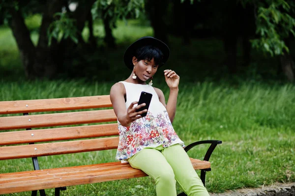 Yeşil pantolonlu, siyah şapkalı, parktaki bankta poz veren ve cep telefonuyla selfie çeken Afro-Amerikan manken kadın..