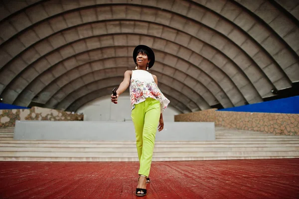 Afro-Amerikan model kadın yeşil pantolon ve siyah şapka şaşırtıcı arena hall karşı açık poz verdi.