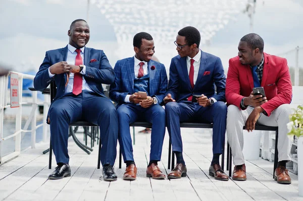 Group of four african american happy succesfull mans at suit. Rich black business mans sitting at chairs looking at mobile phones.