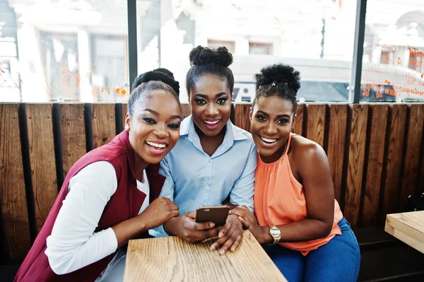 Three african american girls sitting on the table of caffe and looking on mobile phone.