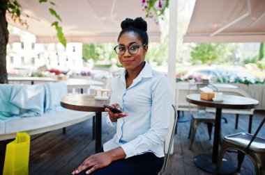 Afro-Amerikan Kız giyim gözlük ile hareket eden telefon açık caffe oturan.