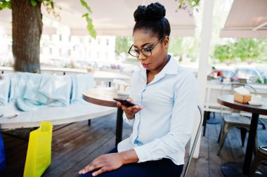 Afro-Amerikan Kız giyim gözlük ile hareket eden telefon açık caffe oturan.