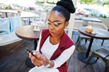 Afro-Amerikan Kız giyim gözlük ile hareket eden telefon açık caffe oturan.